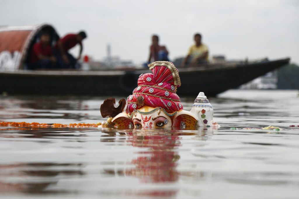 A colorful Ganesha idol partially submerged in a river during a religious ritual, with people on a boat in the background.
