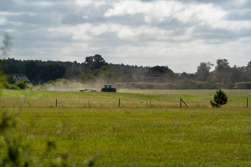 A tractor plowing in a rural field during summer with dust rising, under a cloudy sky.